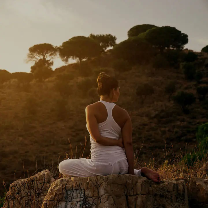 A woman sitting cross-legged on a rock on a hillside at sunset